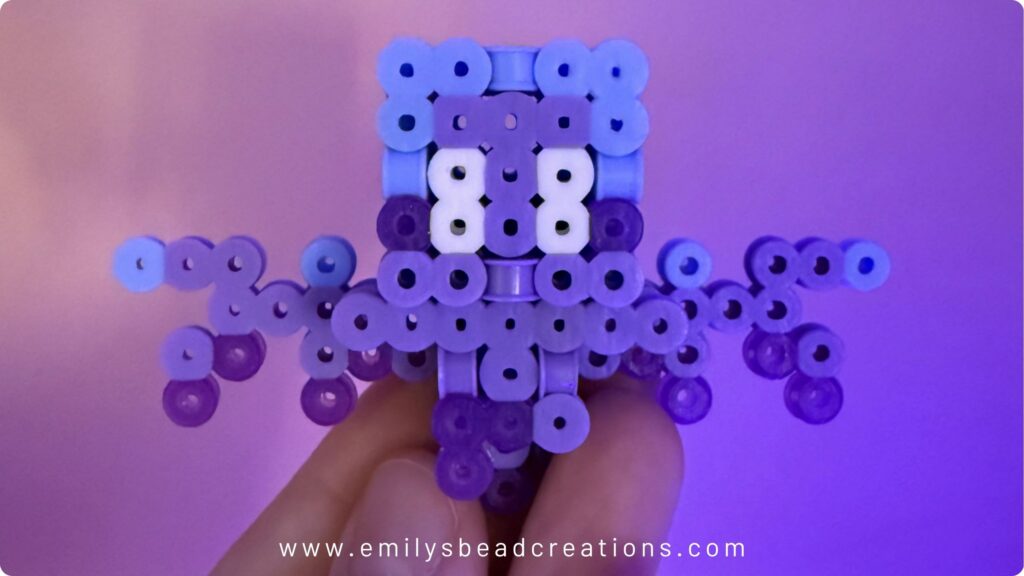 Fingers holding up a 3D Minecraft Allay made out of purple shades of Perler beads rather than the traditional blue.