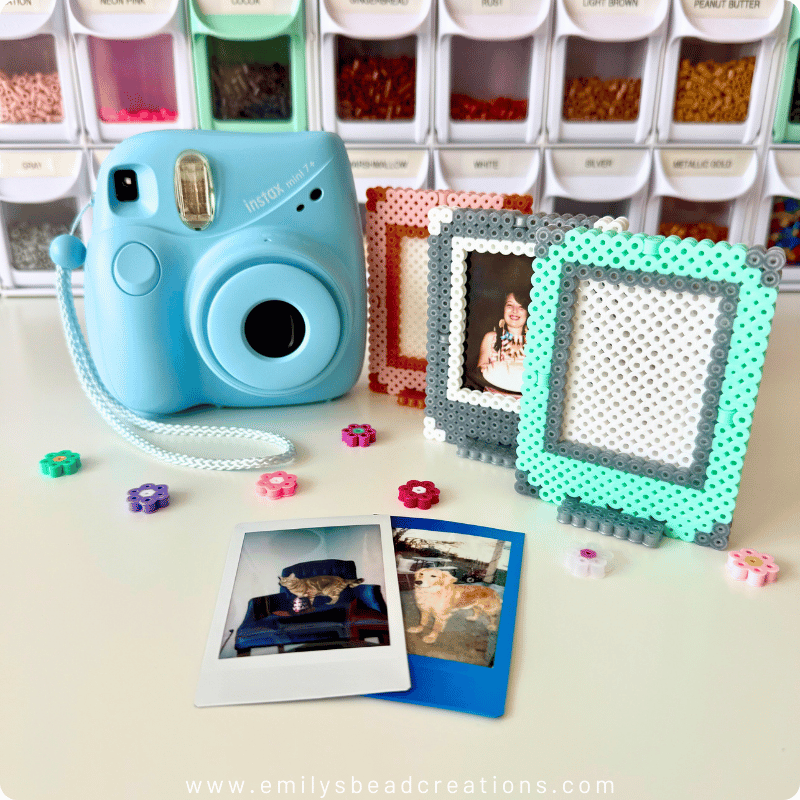 A photo of a baby blue Instax camera beside 3 Perler photo frames (pink, silver, and mint), 2 instant photos in the foreground, and several Perler bead flowers scattered on the white surface. Bins with colorful beads make up half the background.