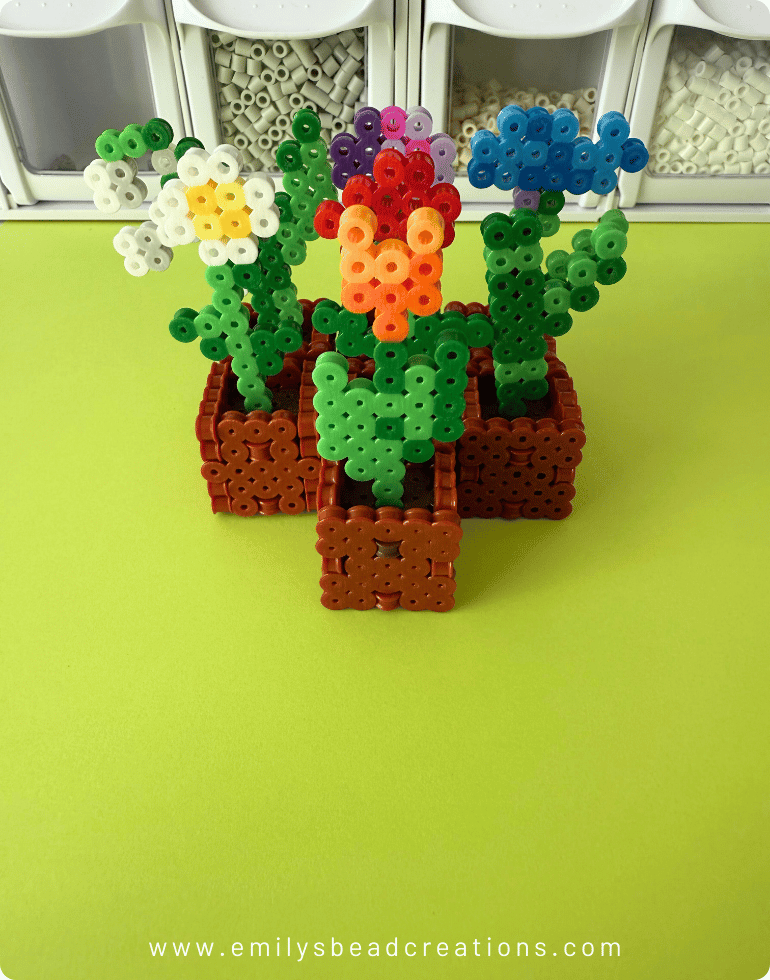 A photo of 7 Minecraft Flower Pots 3D Perler bead creations close together on top of a green surface with bins of Perler beads in the background.